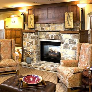 A cozy living room with a stone fireplace, flanked by two patterned armchairs and warm wood paneling on the walls.