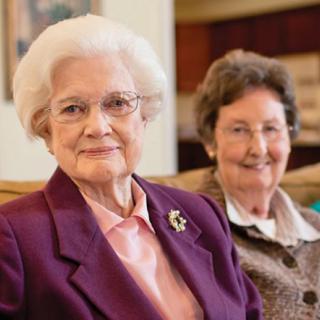Two elderly women sitting together indoors, smiling.