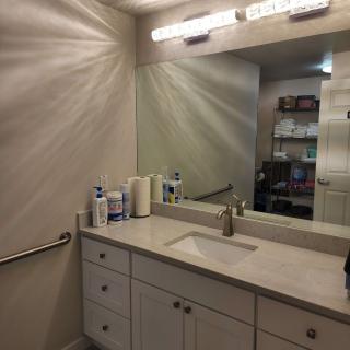 A clean bathroom with a large mirror, countertop sink, and cabinets beneath. It has overhead lighting and various hygiene products on the counter.