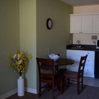 A small dining area with a table and chairs, adjacent to a kitchen, featuring a vase with flowers and a wall clock.