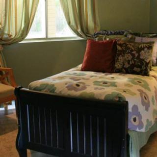 A cozy bedroom with a bed featuring floral bedding and decorative pillows, next to a window with sheer green curtains.