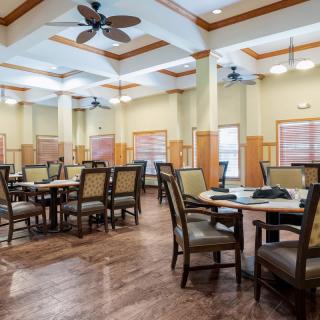 An empty dining hall with neatly arranged tables and chairs, featuring ceiling fans and a wooden floor.