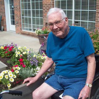 An elderly man sitting by a garden full of colorful flowers.