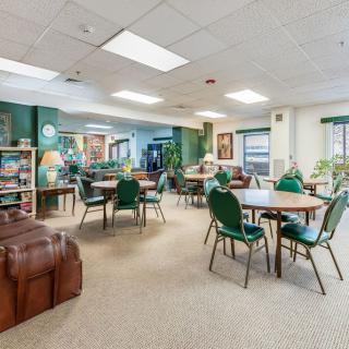 A spacious community room with tables and chairs, decorated in green tones, featuring indoor plants and shelves.