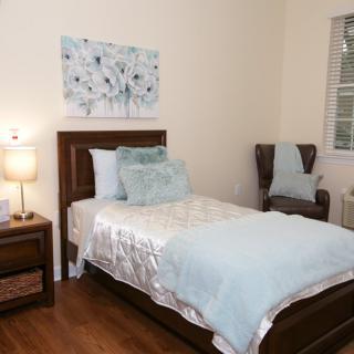 A cozy bedroom featuring a single bed adorned with soft blue and white bedding, complemented by a lamp and wall art.