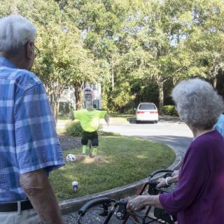 An elderly couple watches a person in reflective clothing working in a neighborhood garden.