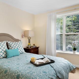 A cozy bedroom with a neatly made bed, decorative pillows, and a bedside lamp, featuring a large window that lets in natural light.
