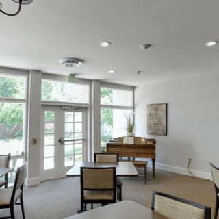 A bright room with a piano, tables, and chairs, illuminated by natural light from large windows.