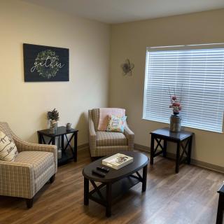 A cozy living room with armchairs, a coffee table, and side tables, featuring wooden floors and natural light from a large window.