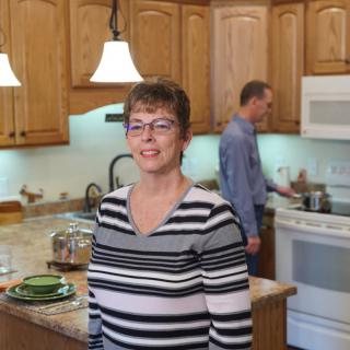A woman smiling in the kitchen while a man is cooking in the background.