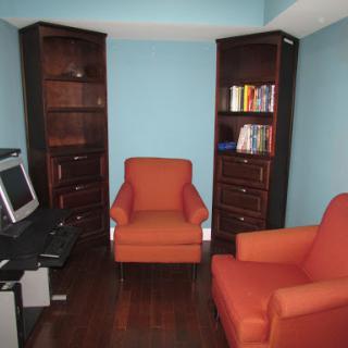 A cozy office space featuring two orange chairs, dark wooden bookshelves filled with books, and a computer setup.
