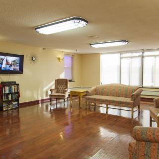 A cozy living room with wooden flooring, featuring a sofa set, chairs, a television mounted on the wall, and a bookshelf.