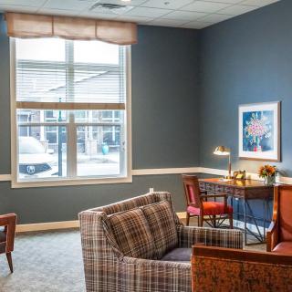 A cozy living room with plaid armchairs, a wooden desk, and large windows letting in natural light.