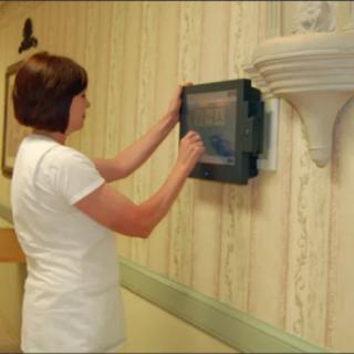 A nurse interacting with a touchscreen device mounted on a wall in a healthcare setting.