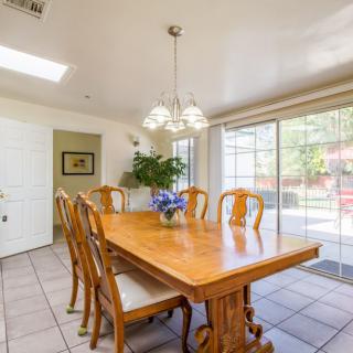 A bright dining room with a wooden table and chairs, illuminated by a chandelier, featuring an indoor plant and a sliding glass door.