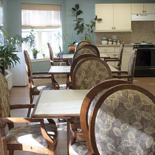 A cozy dining area with elegant chairs and tables, illuminated by daylight from the windows, featuring a kitchen in the background.