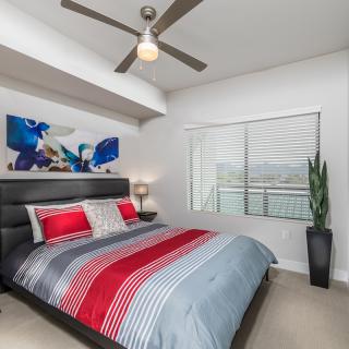 A modern bedroom featuring striped bedding, artwork on the wall, a ceiling fan, and an indoor plant, illuminated by natural light from a large window.