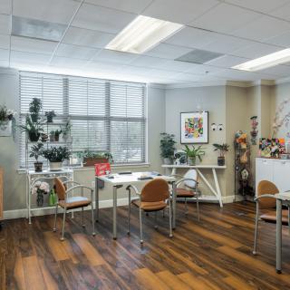 A modern office interior with large windows, decorated with plants and featuring desks and chairs on a wooden floor.