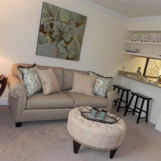 A cozy living room with a beige sofa and an ottoman, accented by a piece of artwork on the wall and bar stools in the corner.