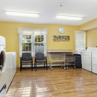 A clean and spacious laundry room with yellow walls, featuring a washer, dryer, chairs, and wooden flooring.
