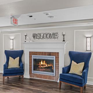A cozy living room with a fireplace, featuring blue chairs and a 'Welcome' sign.