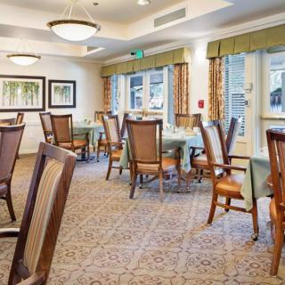 A well-lit dining room with tables and chairs arranged neatly, featuring large windows and carpeted flooring.