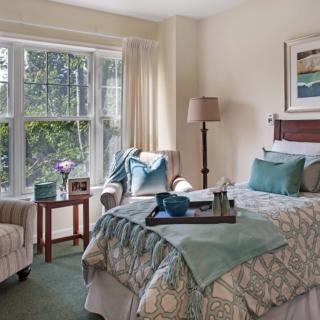 A cozy bedroom with soft lighting and comfortable furnishings, featuring a bed, a chair by a large window, and tasteful decor.