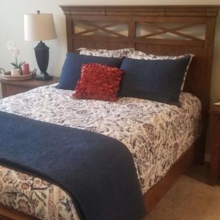 A cozy bedroom with a bed featuring blue and patterned bedding, a decorative red pillow, and a wooden headboard. A lamp and nightstand are positioned beside the bed.