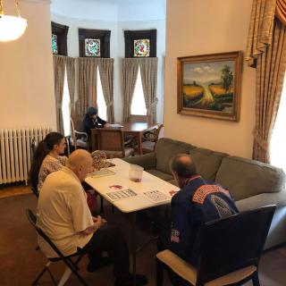 A group of people sitting at a table having a discussion in a well-lit room with curtains and a painting on the wall.