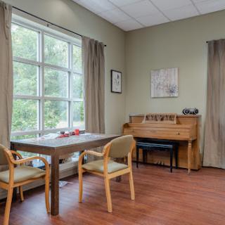 A cozy and well-lit interior featuring a wooden table and chairs, a piano, and large windows bringing in natural light.