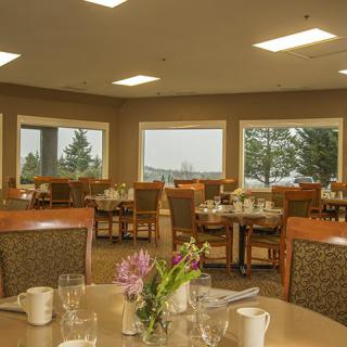 A well-lit dining room with neatly arranged tables and chairs, featuring floral centerpieces and large windows offering a scenic view.