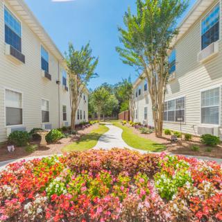 A beautifully landscaped apartment complex with colorful flowers and a central pathway under a clear blue sky.