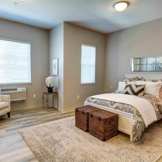 A modern bedroom with neutral tones, featuring a comfortable bed, a wooden chest at the foot of the bed, and natural light streaming in through two windows.