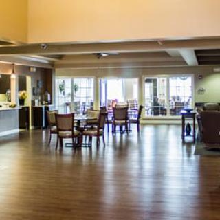 A spacious dining area with wooden flooring and several tables and chairs, well-lit by natural light from large windows.