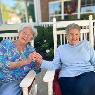 Two elderly women enjoying a moment together while sitting on rocking chairs outside.