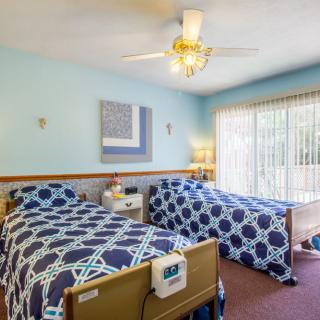 A well-lit nursing home bedroom with twin beds, patterned bedding, and a ceiling fan.