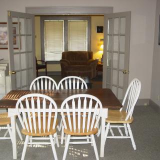 A dining area with a wooden table and white chairs, leading to a living room through French doors.