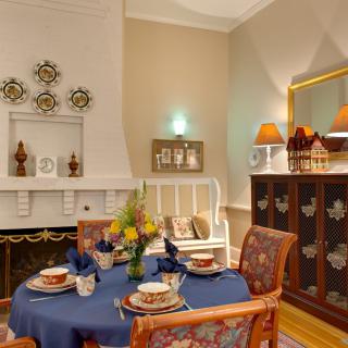 A warm and inviting dining room featuring a round table with a blue tablecloth, surrounded by patterned chairs. The decor includes elegant wall art, a decorative cabinet with lamps, and a cozy fireplace.