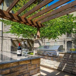 A sunny outdoor kitchen with a grill under a wooden pergola, surrounded by a stone counter and shaded by trees.