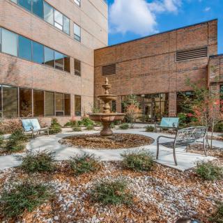 A peaceful courtyard with a central fountain, surrounded by benches and chairs, adjacent to a modern building.