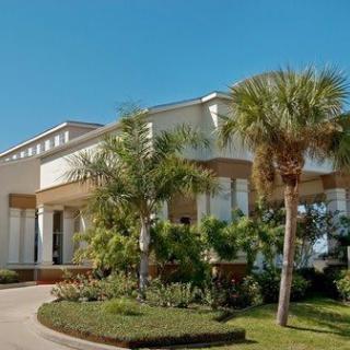 A modern residential house surrounded by palm trees and well-maintained landscaping under a clear blue sky.