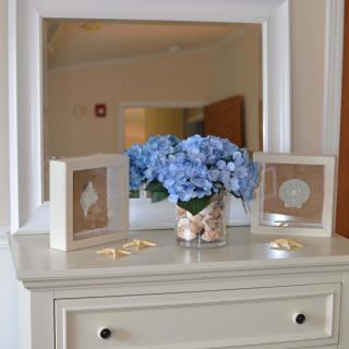 A well-decorated dresser featuring a vase of blue hydrangeas, framed art pieces, and an elegant mirror.