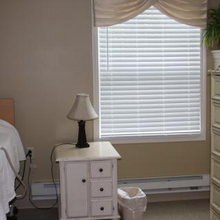 A simple bedroom with a nightstand and lamp next to a bed, featuring a window with blinds and curtains.