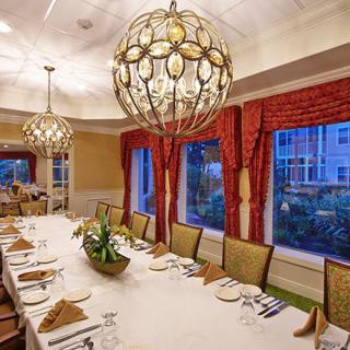 An elegantly decorated dining room with a long table set for a formal gathering, featuring large chandeliers and red curtains framing the windows.