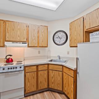 A compact kitchen with wood cabinets, featuring an oven, refrigerator, sink, and a wall clock.