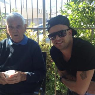 A smiling man with sunglasses and a cap poses next to an elderly person outdoors.