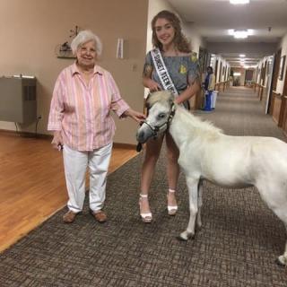A young woman with a sash holds the lead of a miniature horse next to an elderly woman in a hallway.
