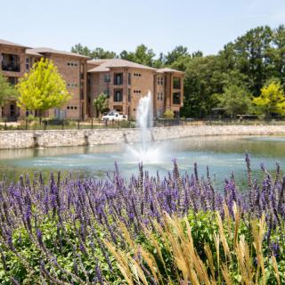 A picturesque view of an apartment complex with a fountain in a pond, surrounded by lush greenery and flowers.
