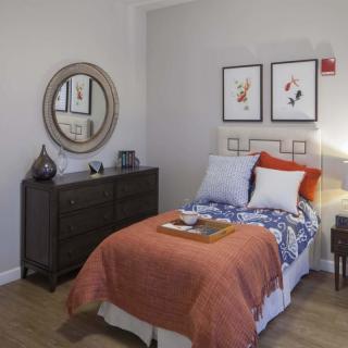 A cozy bedroom with a single bed adorned with colorful pillows and a throw blanket, featuring wall art, a mirror, and a dark wooden dresser.