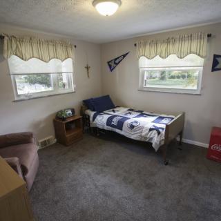 A small bedroom with a single bed, carpeted floor, and two windows adorned with curtains. The room features decorative pennants and wall art.